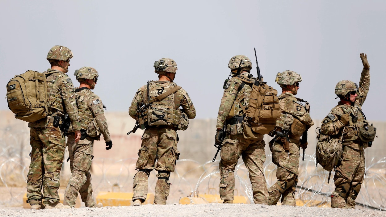 Six soldats américains marchent sur un sol terreux, le long d'une clôture de barbelés.