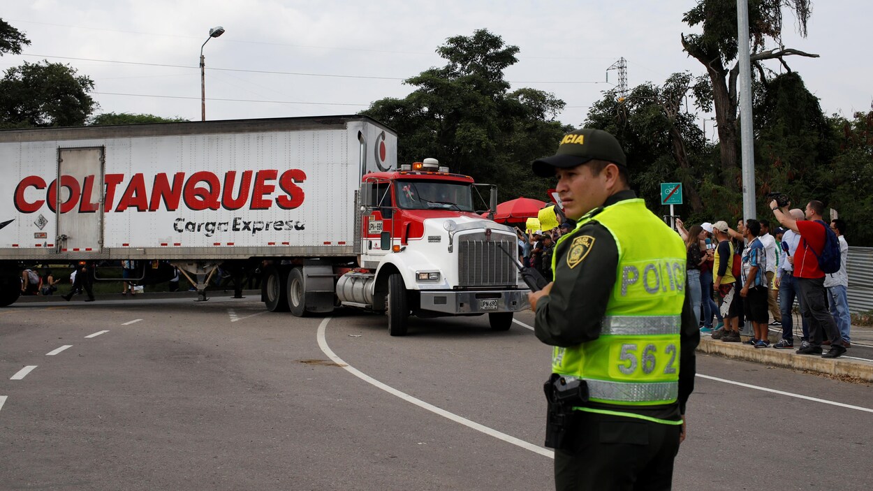 Un policier se tient dans une rue où un camion est stationné. Des gens manifestent tout près du camion.