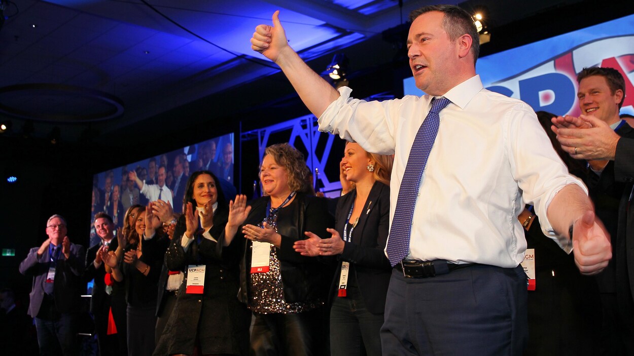Jason Kenney salue ses militants d'un pouce levé, sous les applaudissements de ses ministres, debout derrière lui.