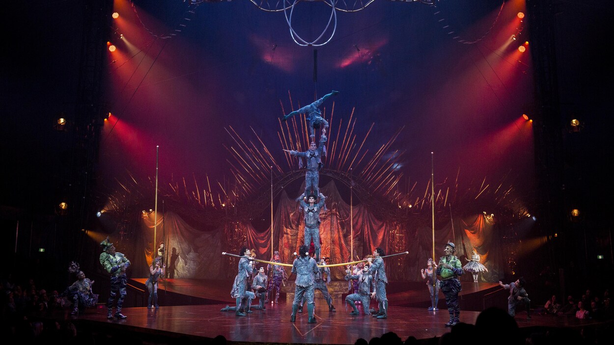 Trois acrobates costumés se tiennent en équilibre sur une perche tenue par d'autres artistes du Cirque du Soleil, pendant que d'autres personnages costumés les observent sur la scène.