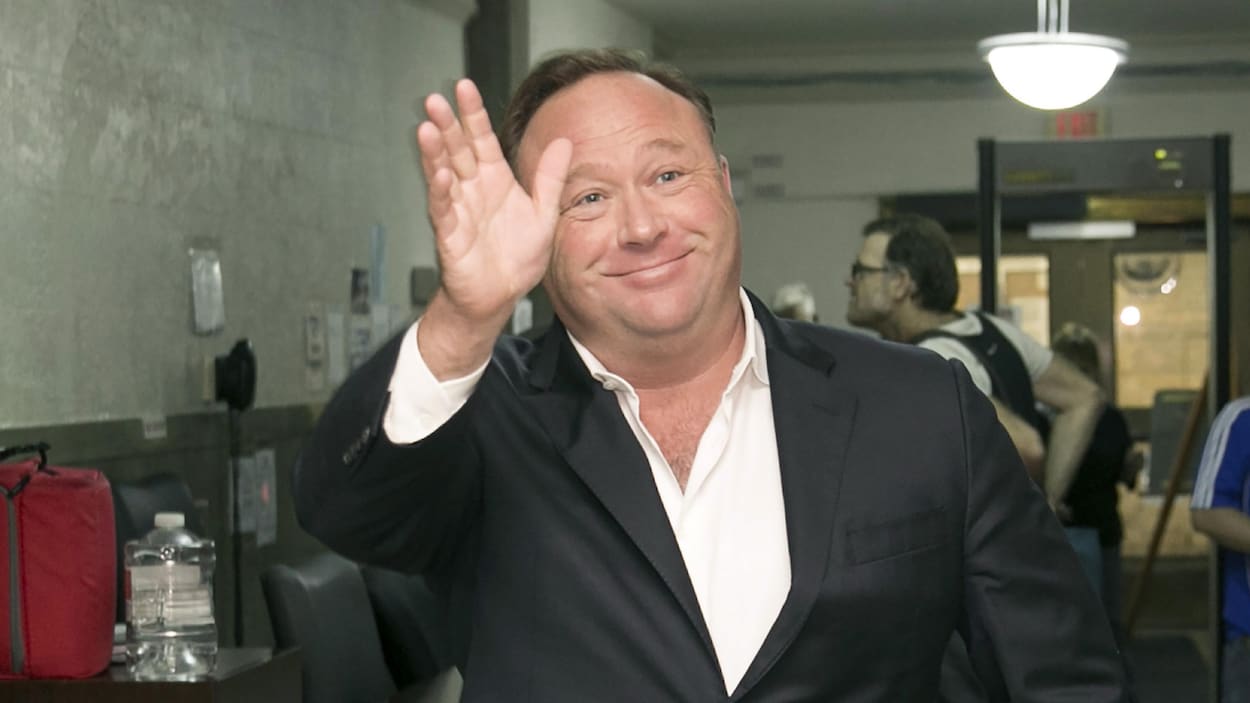 Une photo montrant Alex Jones, souriant, en train de saluer quelqu'un hors du cadre de la caméra. Une photo montrant Alex Jones, souriant, en train de saluer quelqu'un hors du cadre de la caméra.