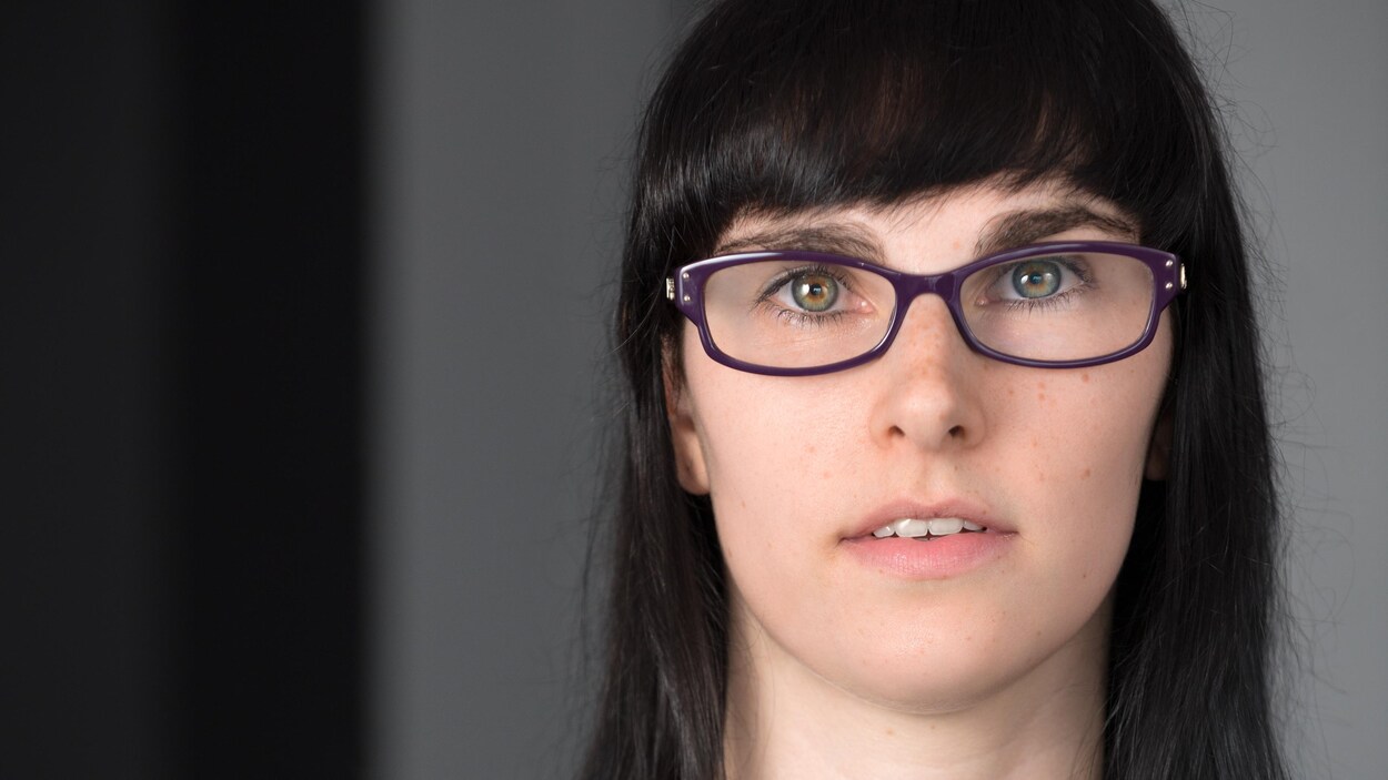 Portrait en couleur de l'auteure Alex Thibodeau sur fond gris : elle porte des lunettes mauves, un chandail gris, des cheveux bruns lâchés et une frange.
