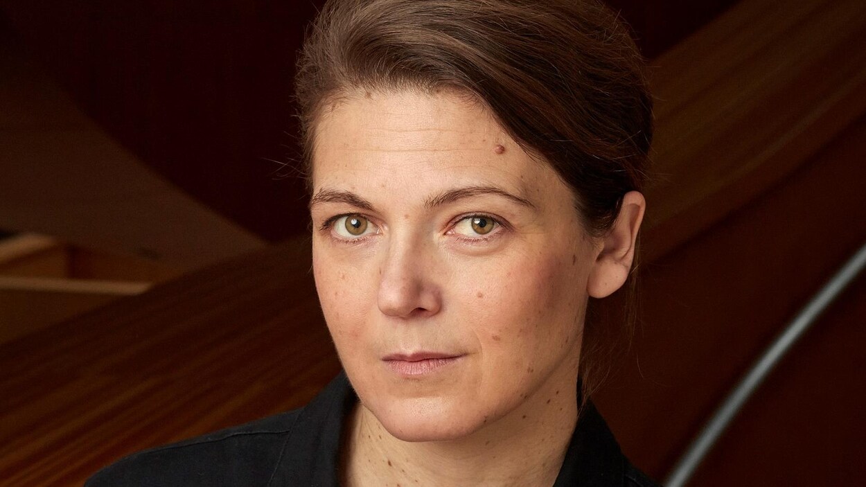 Une femme aux cheveux tirés vers l'arrière fixe la caméra.