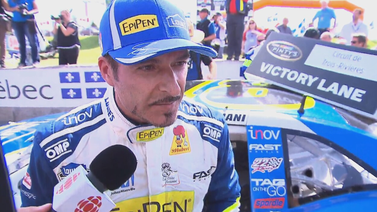 Le pilote Alex Tagliani avec un casquette bleu, devant sa voiture répond aux questions des journalistes