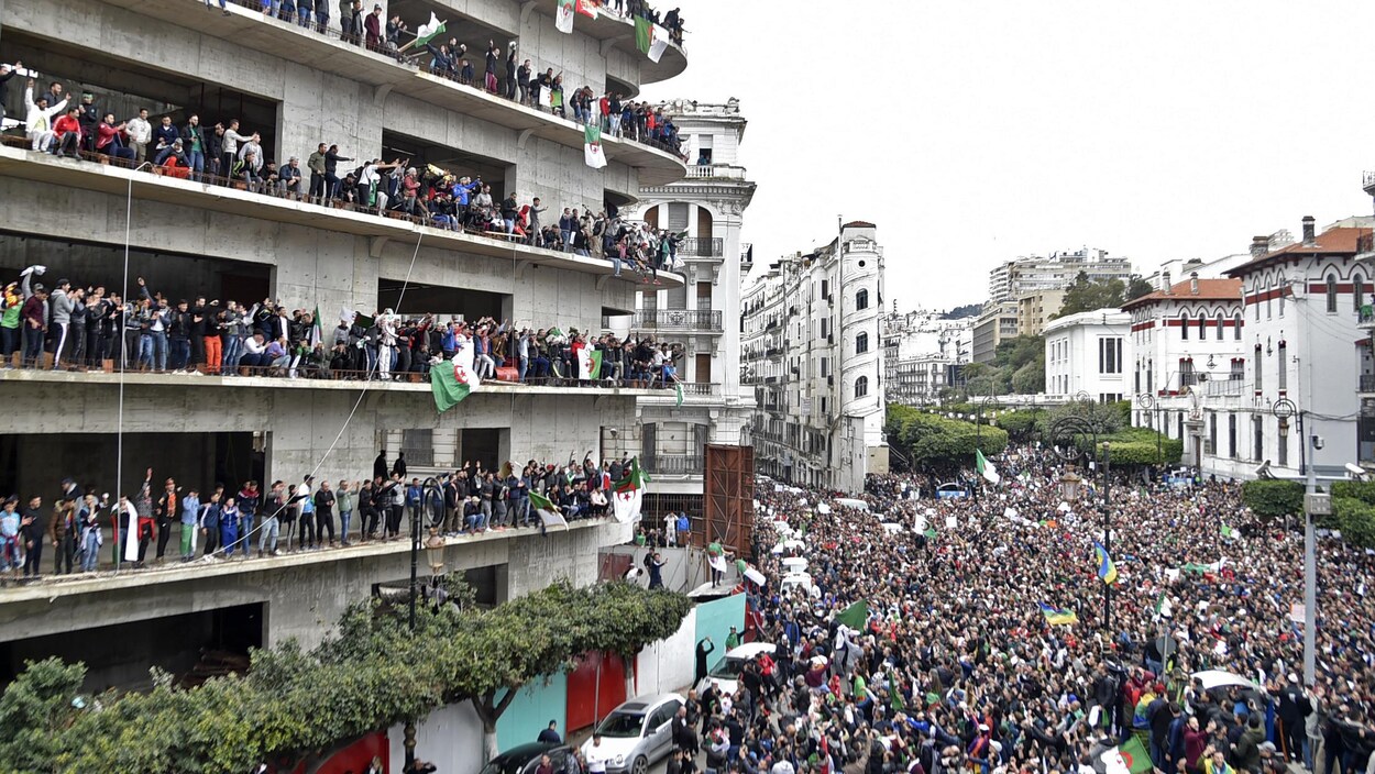 Plusieurs milliers de manifestants marchent dans les rues de la ville alors que d'autres sont debout aux balcons d'un bâtiment.