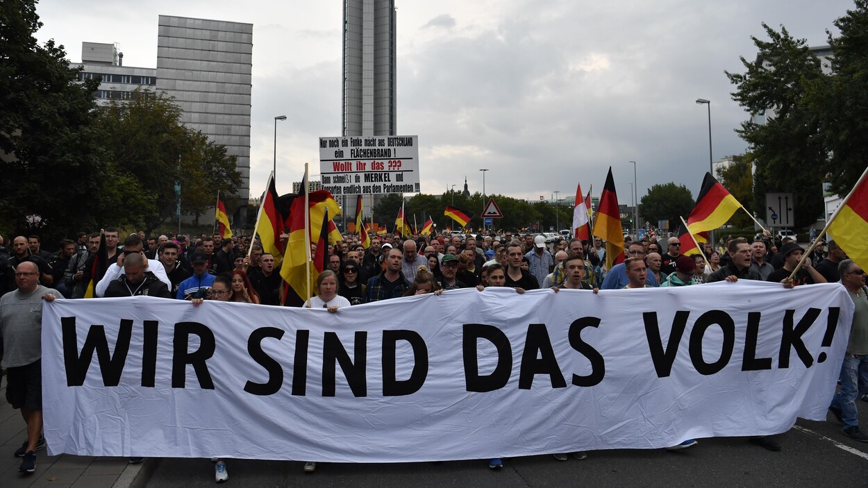 Des Allemands brandissent une longue banderole blanche sur laquelle il est écrit en lettres majuscules: «Nous sommes le peuple» en plus de brandir des drapeaux de l'Allemagne.