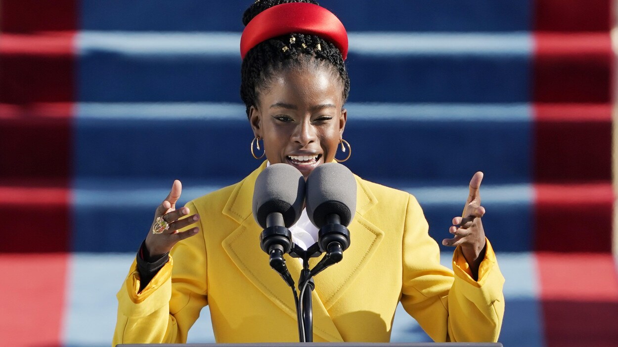 La jeune femme noire porte un veston jaune et un accessoire rouge dans les cheveux. Elle est debout derrière deux micros et lève ses mains aux épaules.