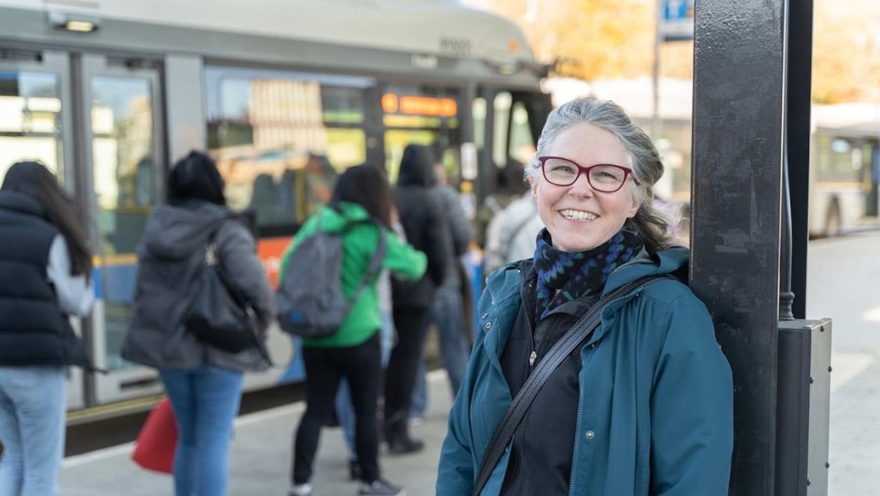 Plan moyen d'Amy Hanser adossée à un poteau avec en arrière-plan des passagers qui montent dans un autobus.
