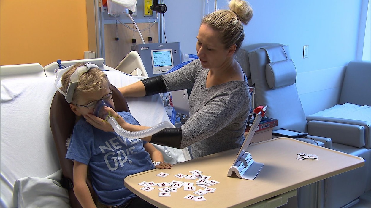 Un enfant est dans un lit d'hôpital, sa mère tient un respirateur sur sa bouche.