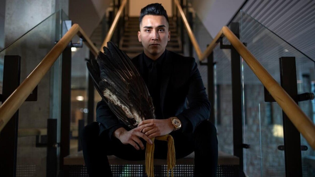 Un jeune homme, Andre Bear, est assis dans un escalier. Il est entièrement vêtu de noir et tient dune parure autochtone de plumes d'oiseaux dans ses mains.
