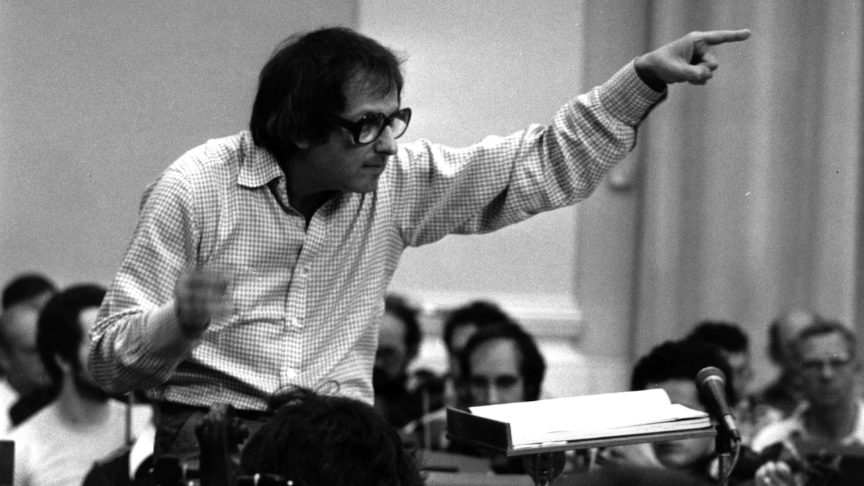 Le musicien André Previn, vêtu d'une chemise à carreaux, a la main gauche levée alors qu'il mène un orchestre au milieu d'une pièce, dans une photo datée de 1979.