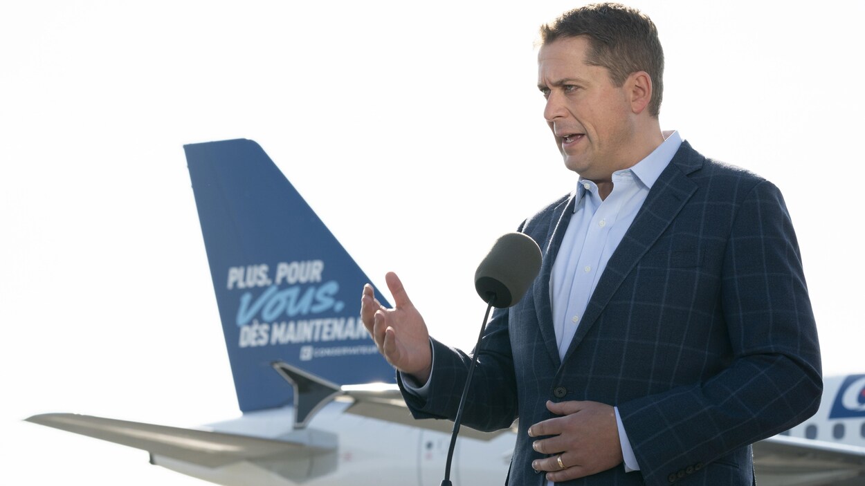 Andrew Scheer, devant la queue d'un avion sur laquelle on peut lire le slogan du Parti conservateur.
