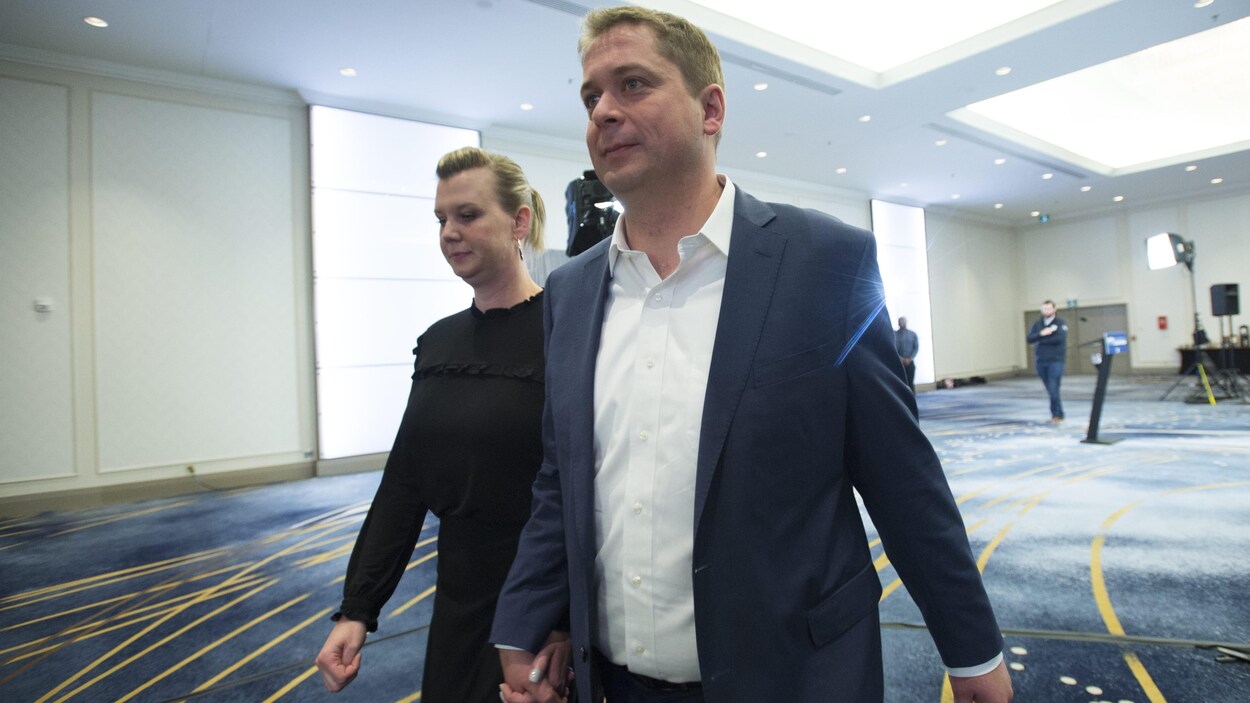 Andrew Scheer et sa femme marchent dans une pièce en se tenant la main.