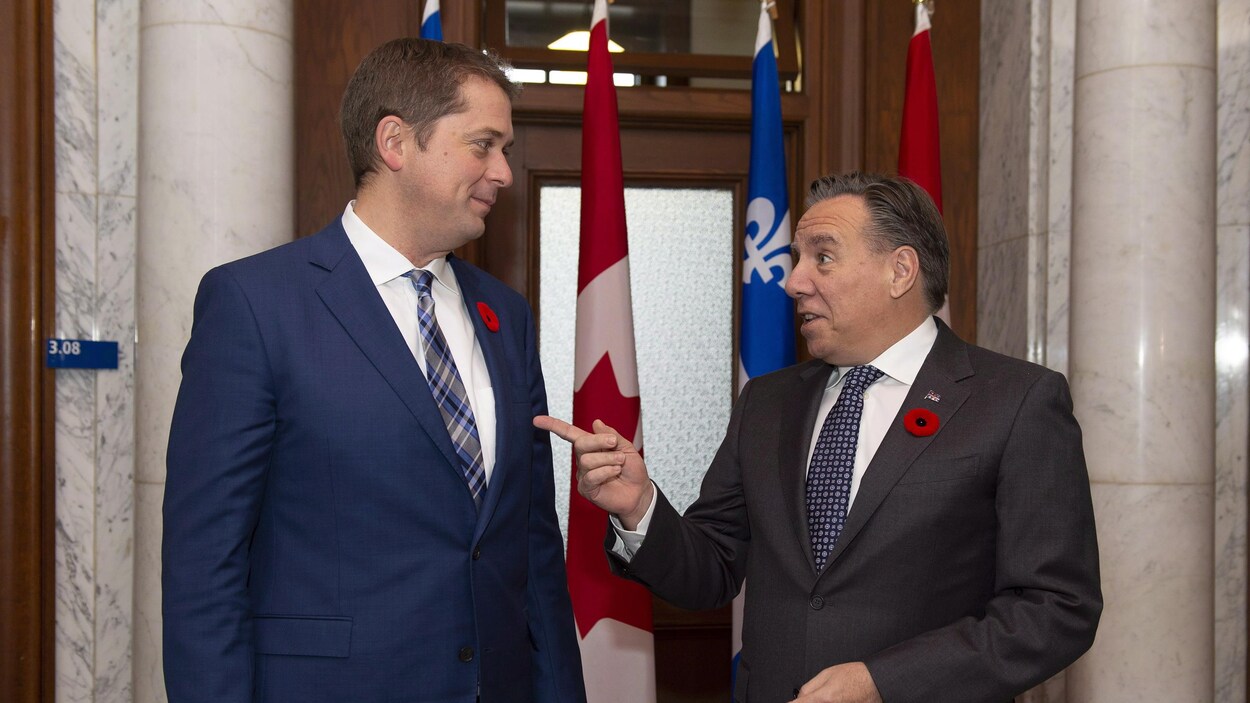Les deux hommes discutent devant des drapeaux du Canada et du Québec.