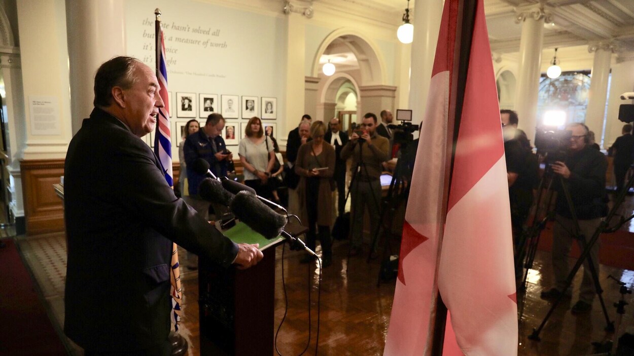 Andrew Weaver debout derrière un lutrin, entre un drapeau canadien et un drapeau de la Colombie-Britannique.