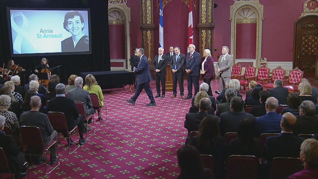 Un groupe de personnes, certains assis, d'autres debout. Derrière eux un écran avec le nom des victimes.