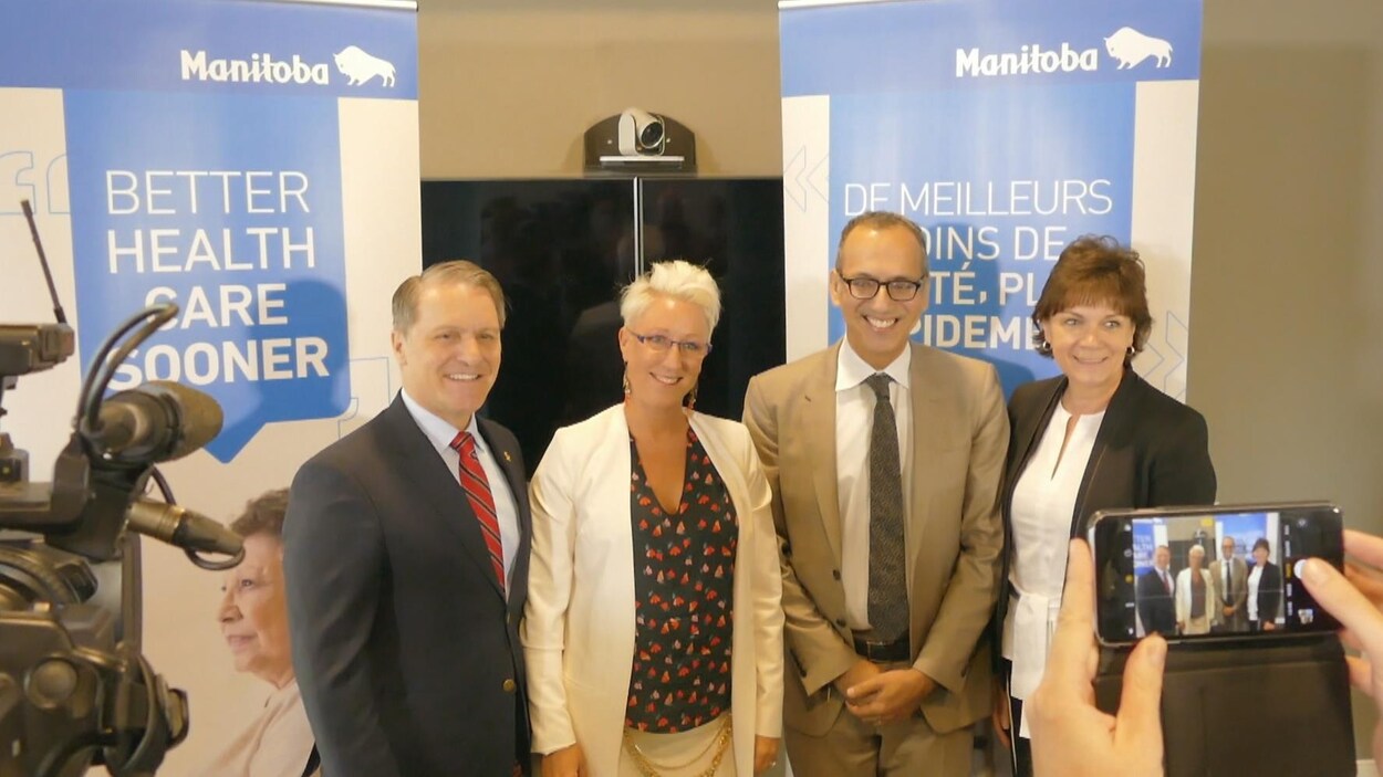 Quatre personnes posent pour une photo devant des panneaux de la province du Manitoba. Au premier plan : des mains tiennent un téléphone qui prend une photo. À gauche, une caméra.