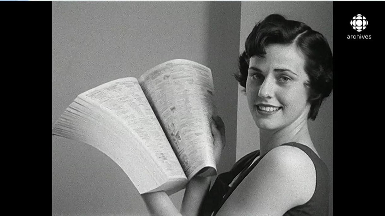 En noir et blanc, femme tout sourire posant avec un bottin téléphoniques ouvert entre ses mains,