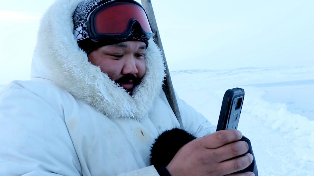 Un homme portant un manteau d'hiver, une tuque et des lunettes de ski regarde son téléphone cellulaire.