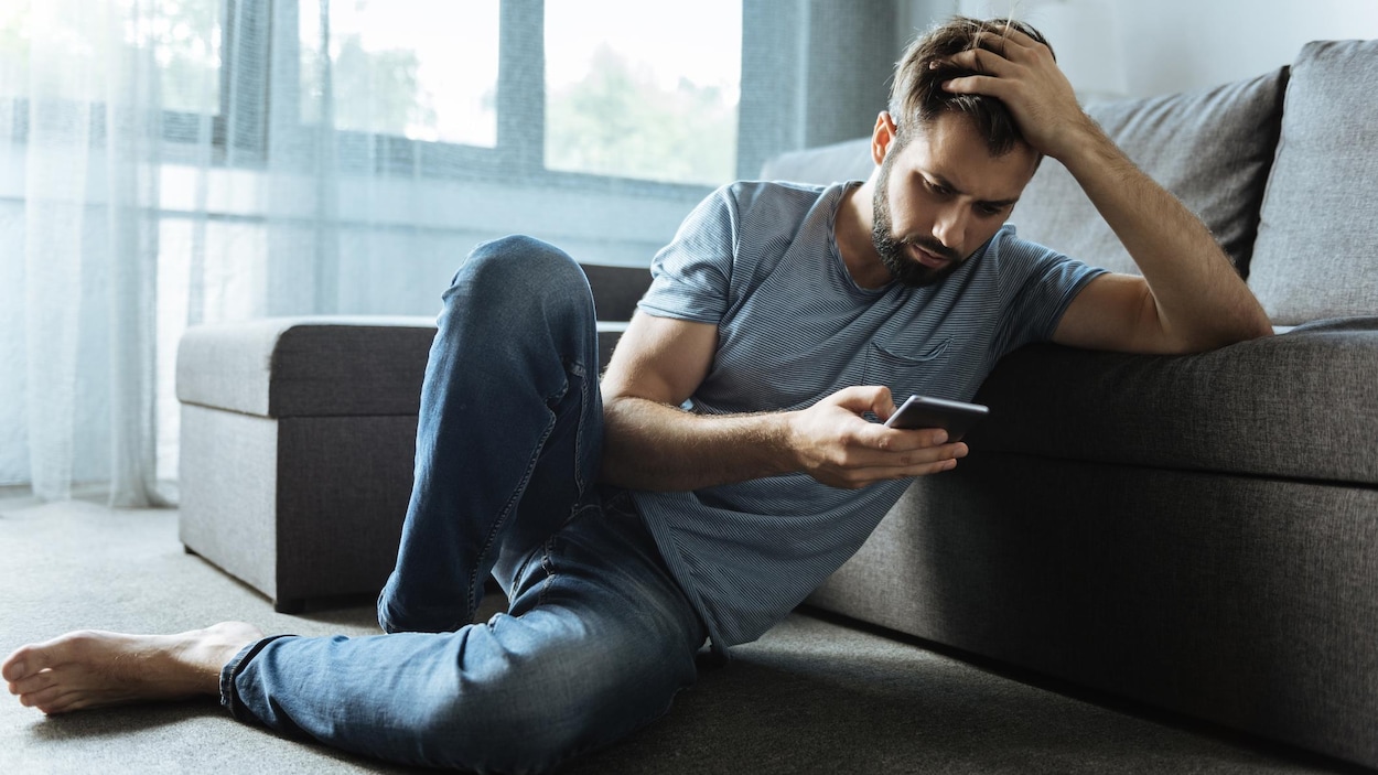 Un homme triste consulte son téléphone. Il est assis sur le tapis de son salon consulte son cellulaire.