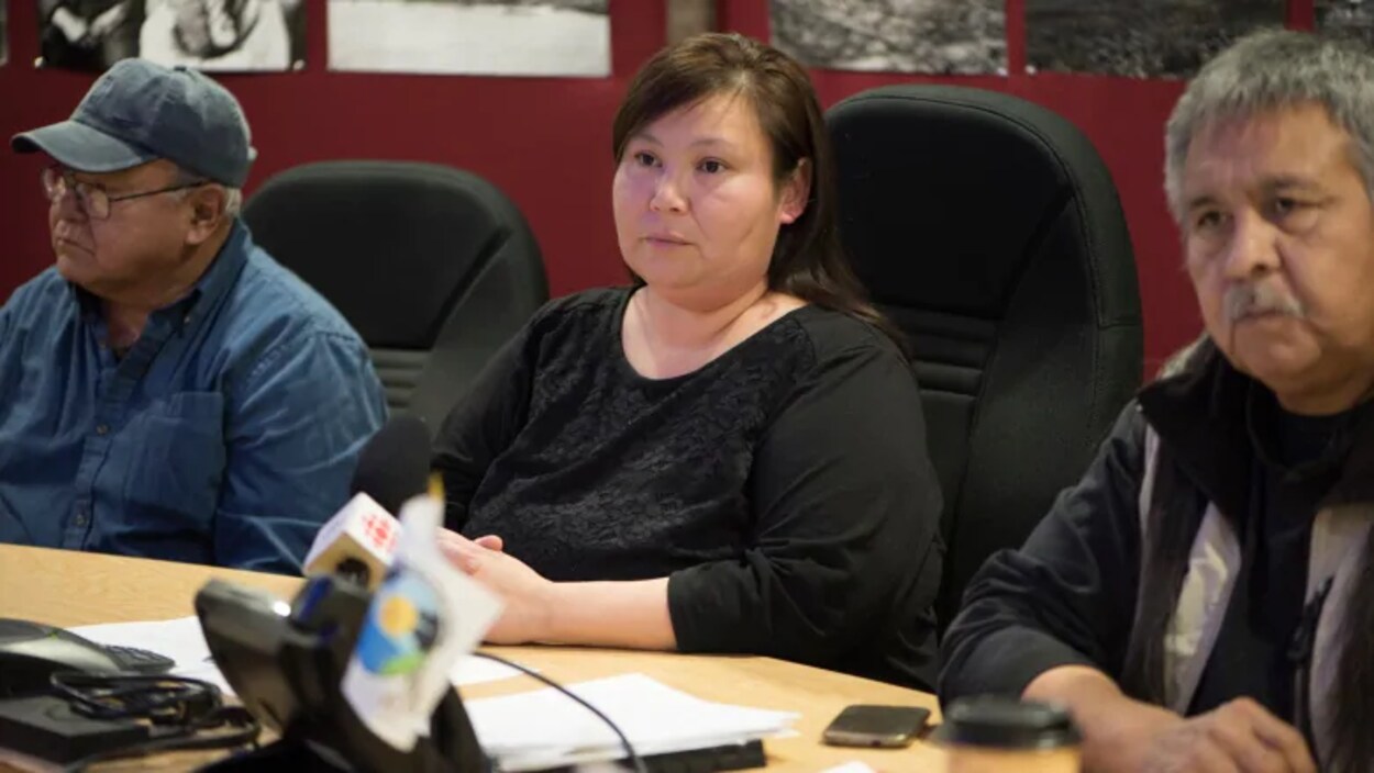 Plan moyen d'April Martel assise entre deux hommes dans une salle de réunion.