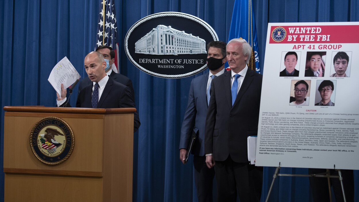 Quatre hommes en complet, dont deux portant un masque. L'un d'eux s'exprime devant une tribune, à côté d'une pancarte montrant la photo de cinq suspects.