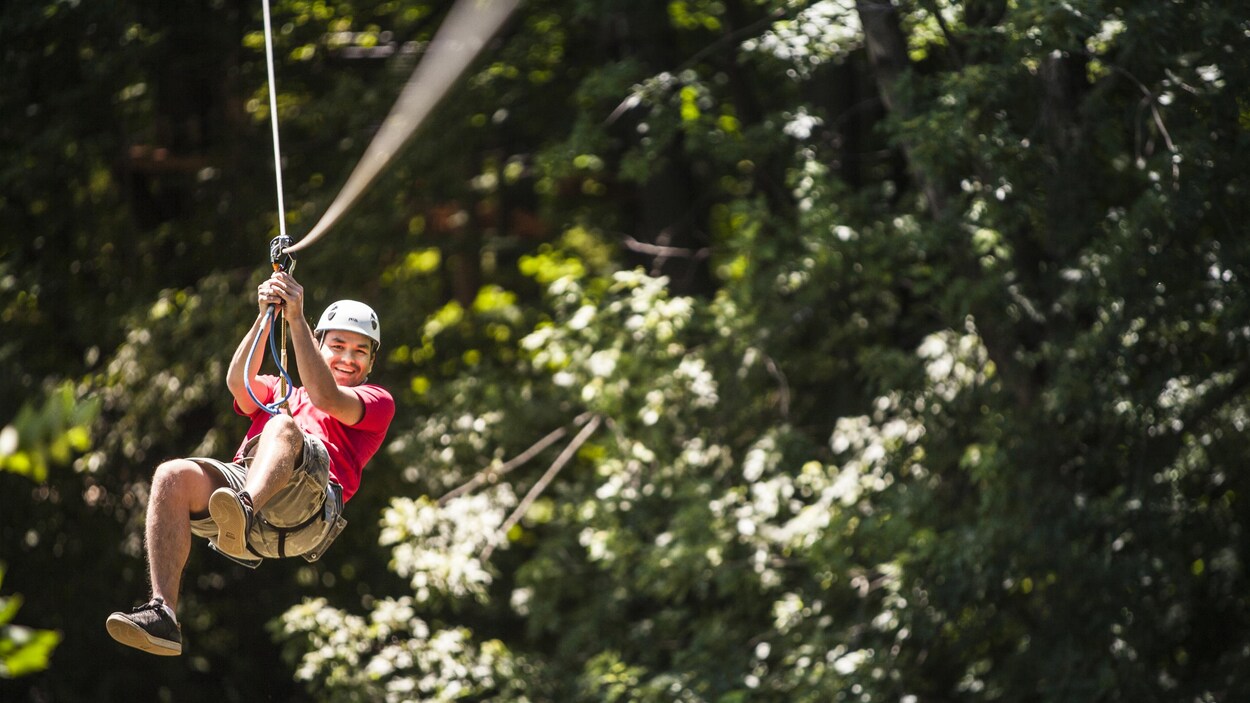 Un jeune homme file, sourire aux lèvres, sur une tyrolienne entre les arbres.