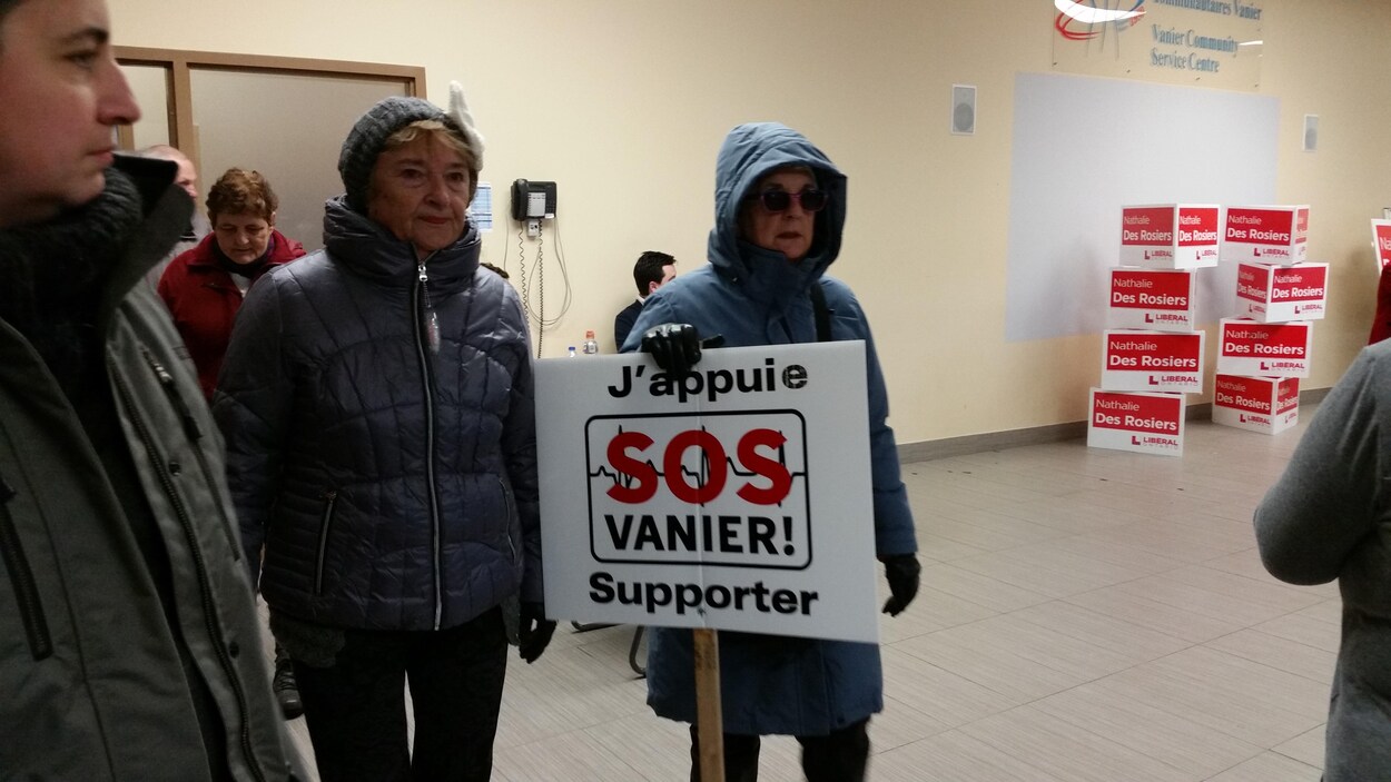 Des gens à l'intérieur d'un centre communautaire. Une femme tient une affiche « J'appuie SOS Vanier ». Des gens à l'intérieur d'un centre communautaire. Une femme tient une affiche « J'appuie SOS Vanier ».