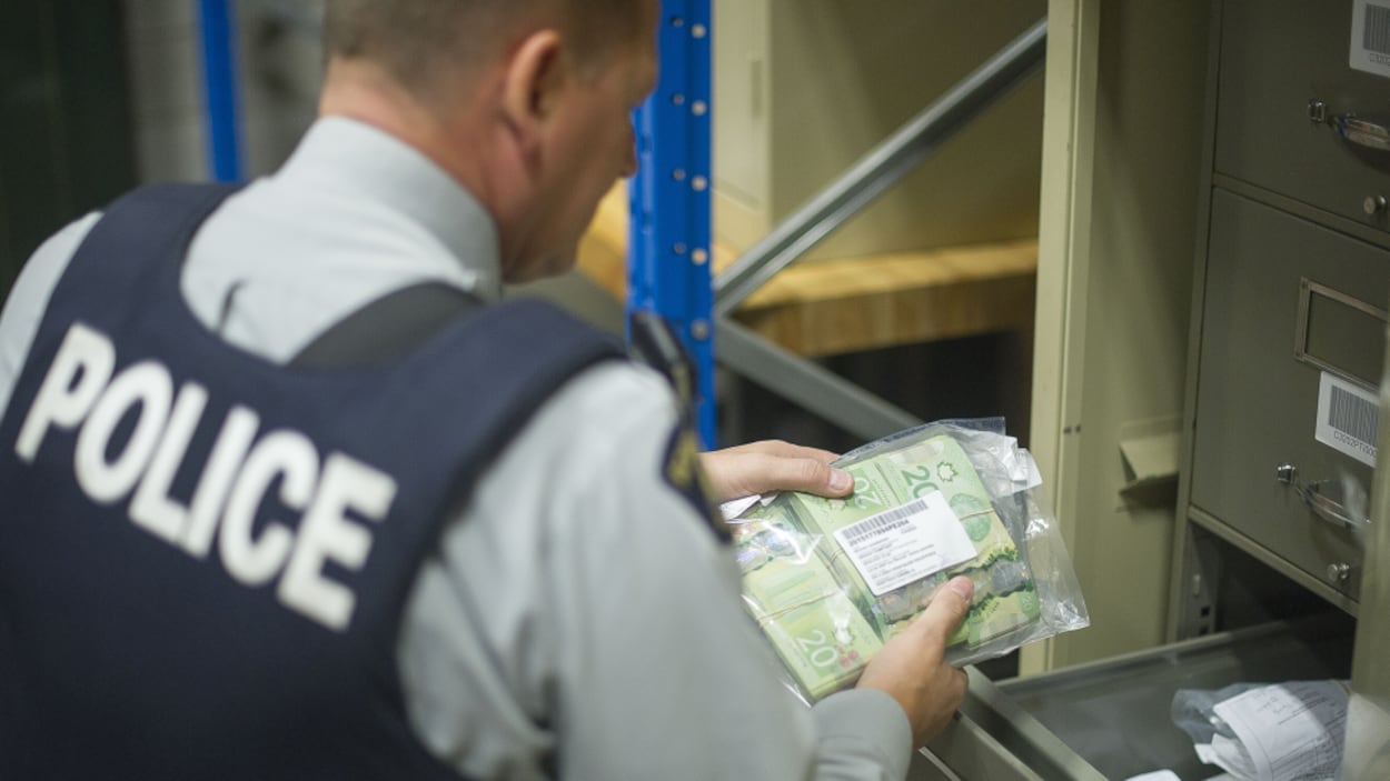 Un policier tient un sac de plastique dans lequel se trouvent plusieurs liasses de billets de 20 dollars. Un policier tient un sac de plastique dans lequel se trouvent plusieurs liasses de billets de 20 dollars.