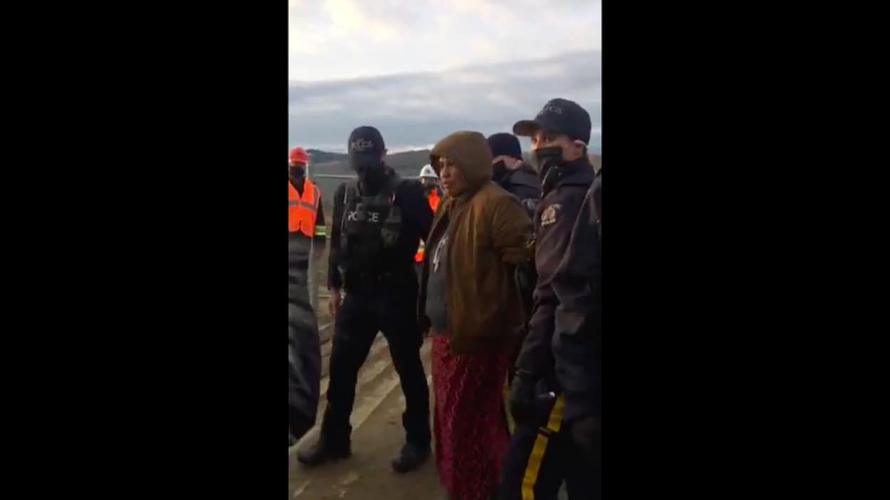 Une femme menottée suivie de 3 policiers avec un travailleur en arrière-plan.