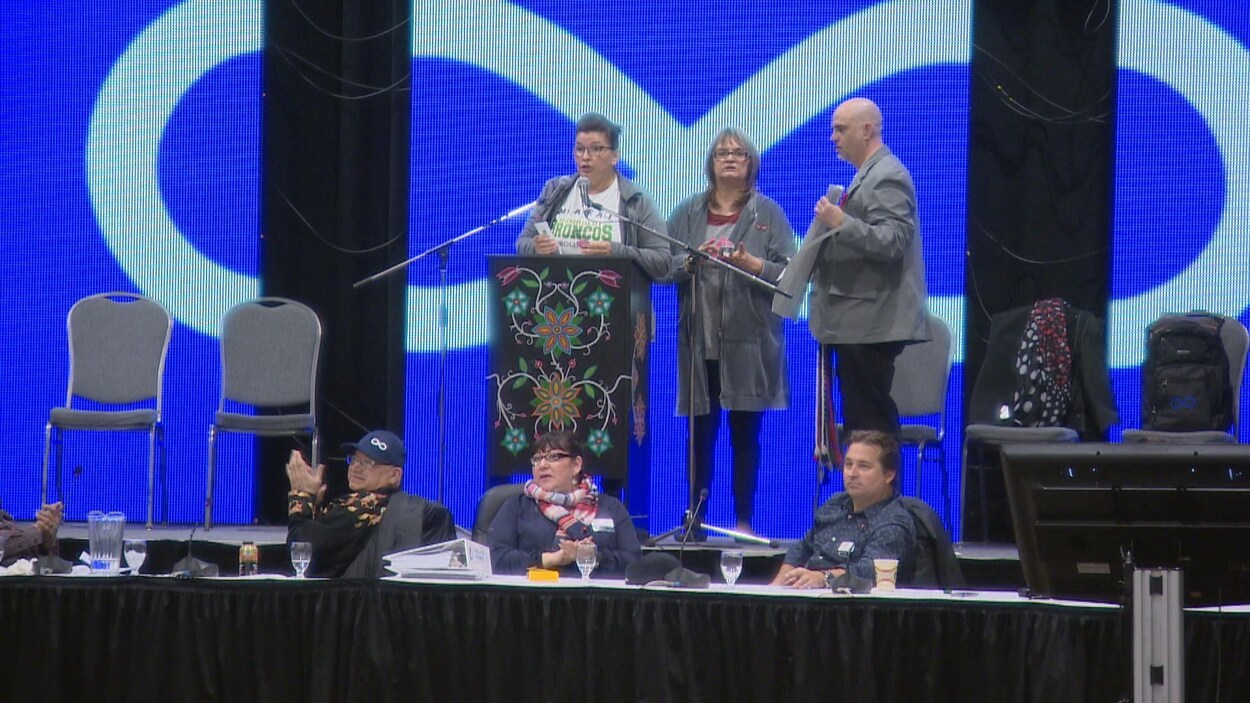Trois personnes sur un podium avec le drapeau métis en arrière-plan. Trois personnes sur un podium avec le drapeau métis en arrière-plan.