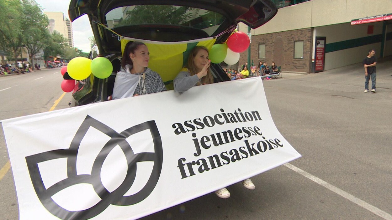 Deux jeunes femmes assises à l’arrière d’un véhicule montrent le logo de l’Asociation jeunesse fransaskoise durant le défilé.