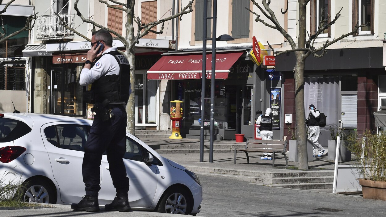 Un policier parle sur un portable à côté d'une voiture stationnée devant des commerces.