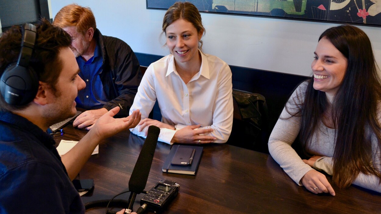 Un journaliste interview deux femmes dans un café. Les trois personnes sont assises autour d'une table et le journaliste tient un micro et porte un casque d'écoute.
