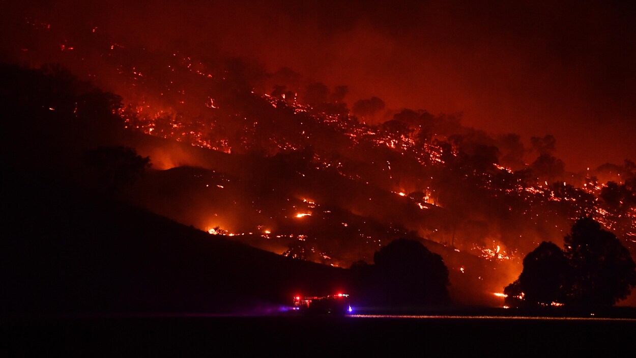 Un camion des pompiers passe devant un flanc de montagne en proie aux flammes dans la nuit.