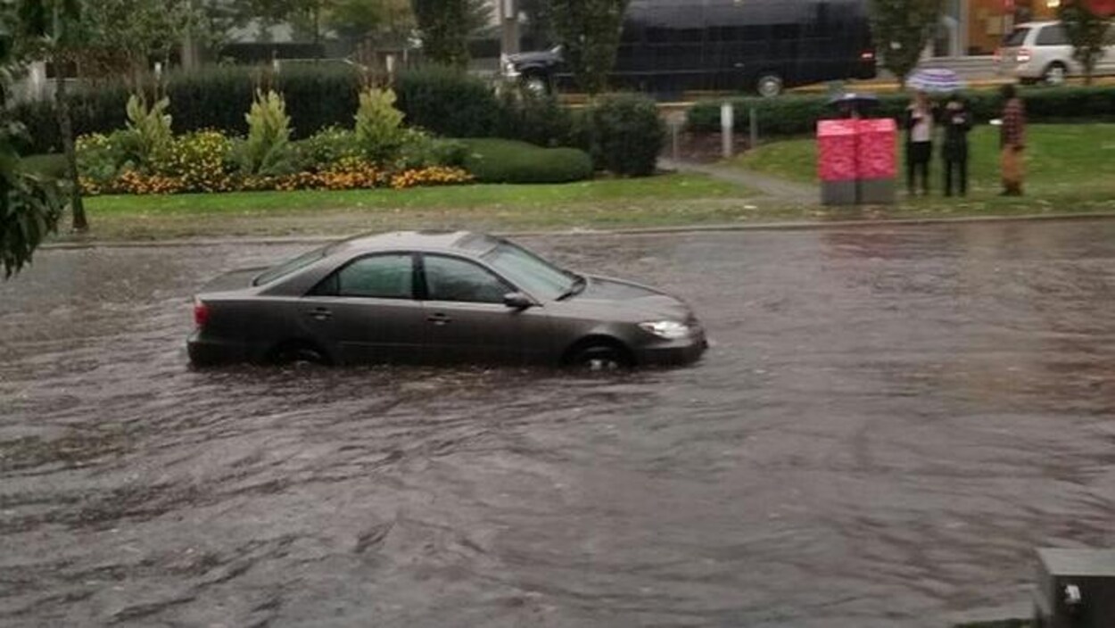 Une voiture en plein milieu d'une rue inondée alors que trois personnes l'observent du trottoir Une voiture en plein milieu d'une rue inondée alors que trois personnes l'observent du trottoir