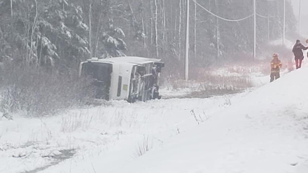 Un autobus renversé sur le côté sur le bord de la route dans la neige avec des pompiers qui se dirigent vers le véhicule.