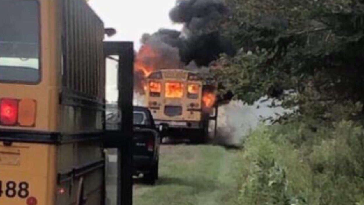 Un autobus scolaire est immobilisé sur le bord de la route et des flammes sont visibles à travers les fenêtres.