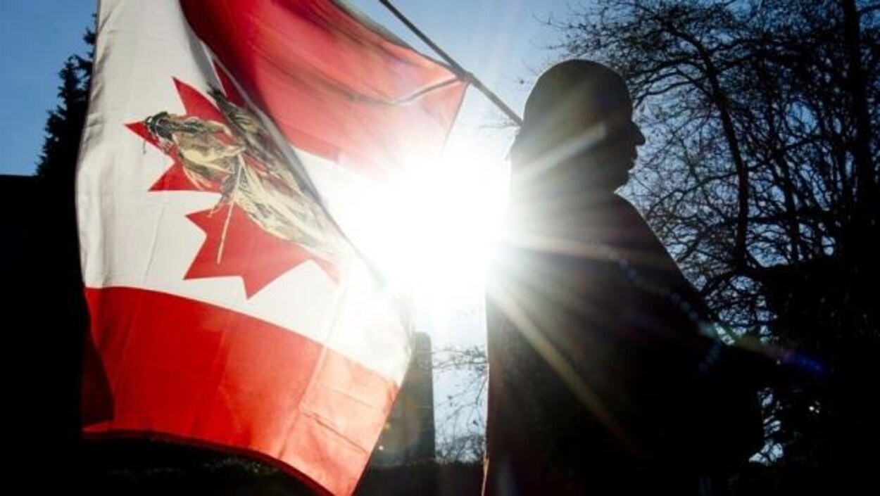 Un membre des Premières Nations tient un drapeau canadien orné d'un dessin de guerrier autochtone. Le cliché est pris en contre-jour. Un membre des Premières Nations tient un drapeau canadien orné d'un dessin de guerrier autochtone. Le cliché est pris en contre-jour.