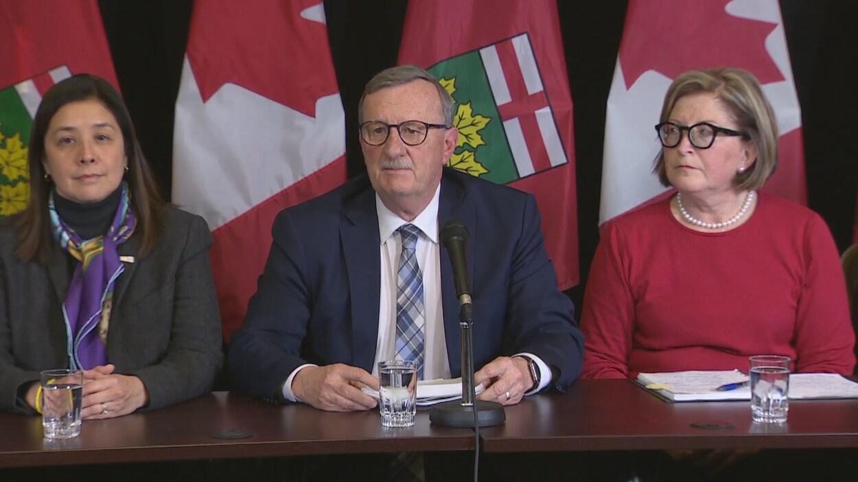 Trois personnes devant des drapeaux dans une salle de presse.