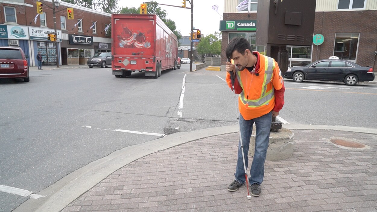Un homme non voyant, portant une veste orange fluorescente, attend au coin d'une rue pour traverser.