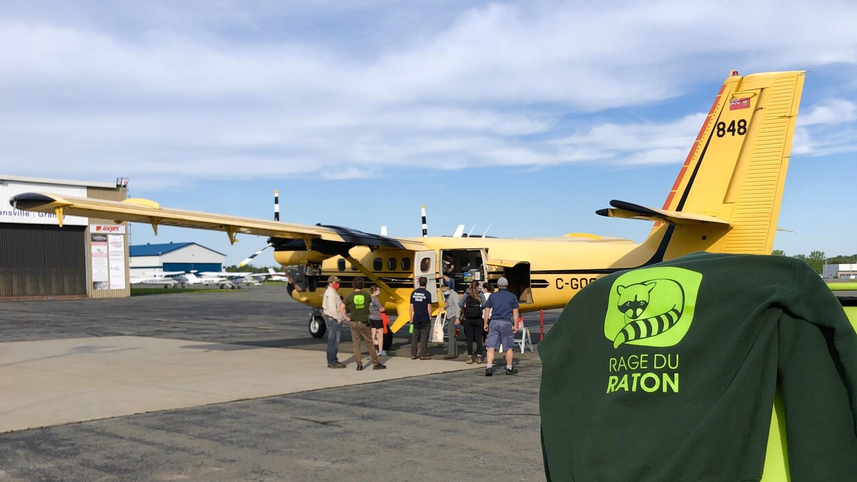 Des gens se tiennent devant un avion en arrière plan et un chandail avec l'indication ''rage du raton'' est en premier plan.