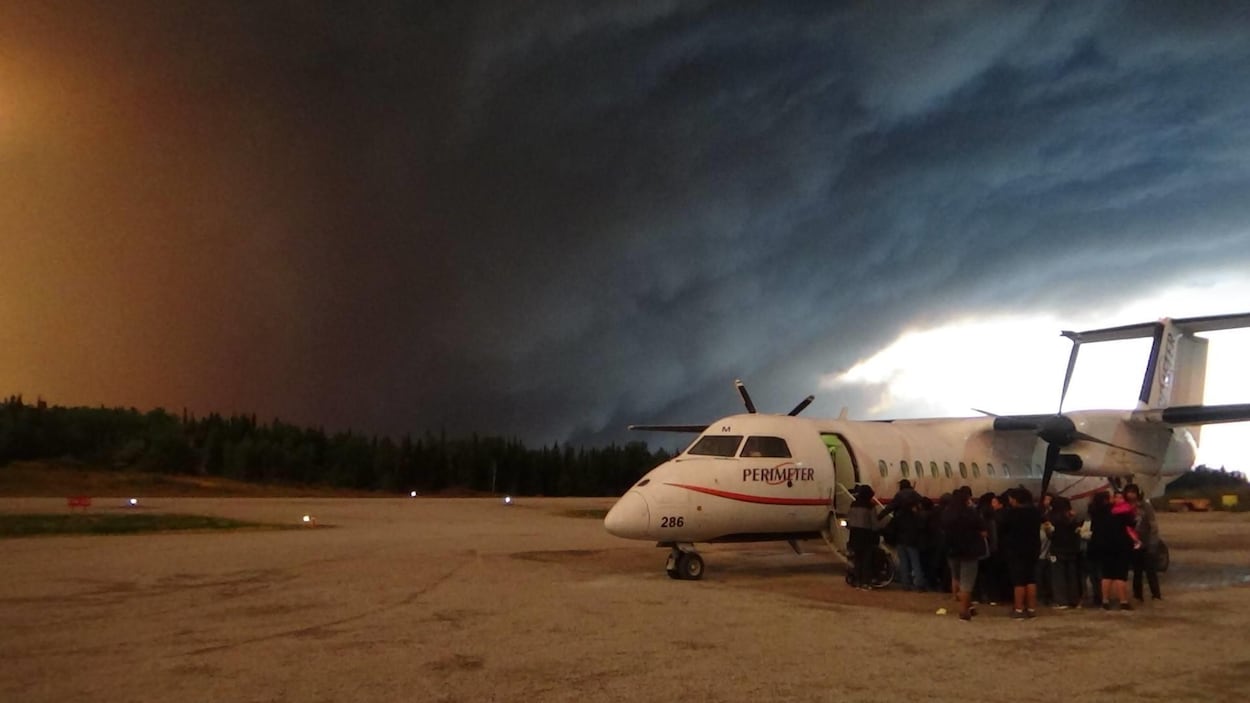 Des personnes font la file pour prendre un petit avion. Il y a une fumée épaisse dans le ciel.