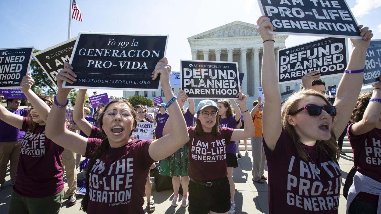 De jeunes femmes portent des t-shirts sur lesquels on peut lire « Je suis la génération pro-vie » manifestent devant la Cour suprême. Certaines portent des pancartes avec le slogan « Cessez de financer les centres de planification familiale ». En arrière-plan, des manifestants pour l'avortement sont présents.