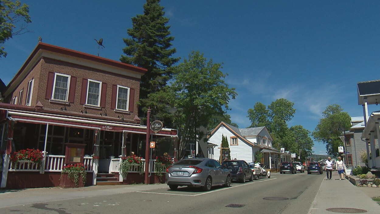 Une rue commerciale avec des voitures stationnées et des marcheurs sur le trottoir, en été, le jour.