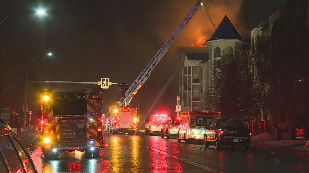 Plusieurs camions de pompiers sont stationnés dans la rue. Un des camions est au milieu de la route, une échelle est déployée et au bout une lance à eau tente d'étendre les flammes qui sortent du dernier étage. Il fait nuit.