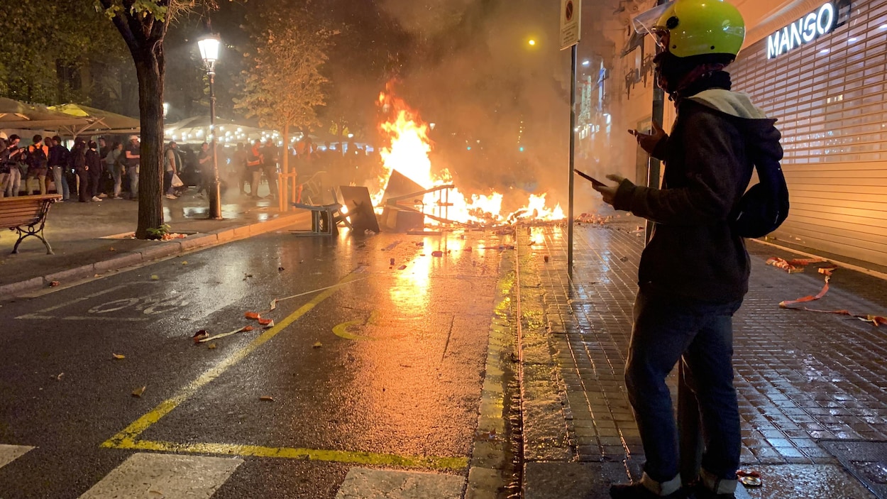 Un homme casqué regarde une foule au loin, une barricade en feu se trouve tout près.
