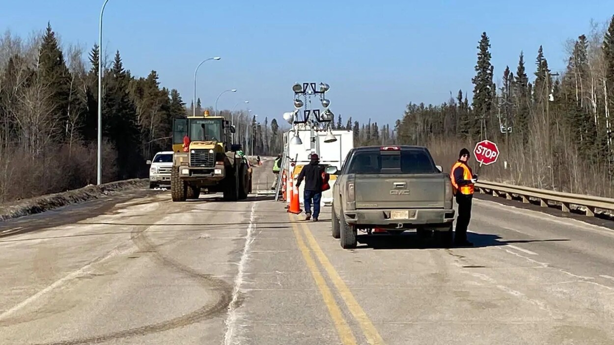 Des véhicules sur la route bloque le passage. Un homme tient un panneau où est écrit stop dans ses mains.