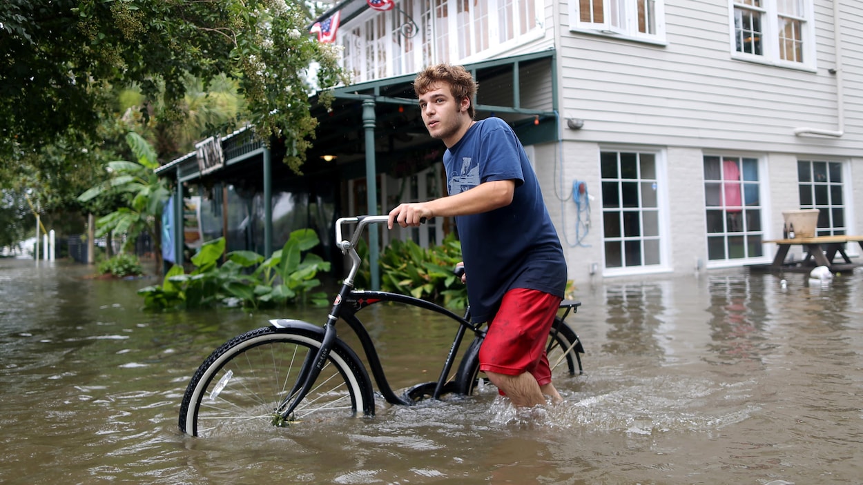 Un jeune homme pousse son vélo dans la rue, avec de l'eau jusqu'aux genoux.