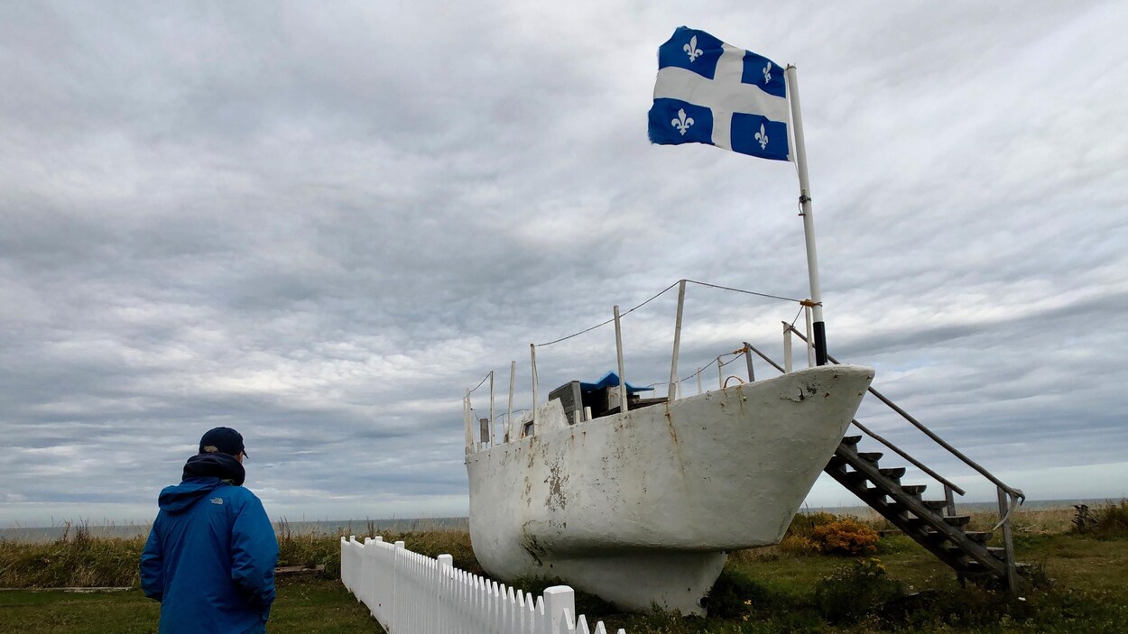 Un homme regarde au large à la hauteur de Pointe-au-Père dans le Bas-Saint-Laurent. À sa droite se trouve un bateau et un drapeau du Québec qui flotte.